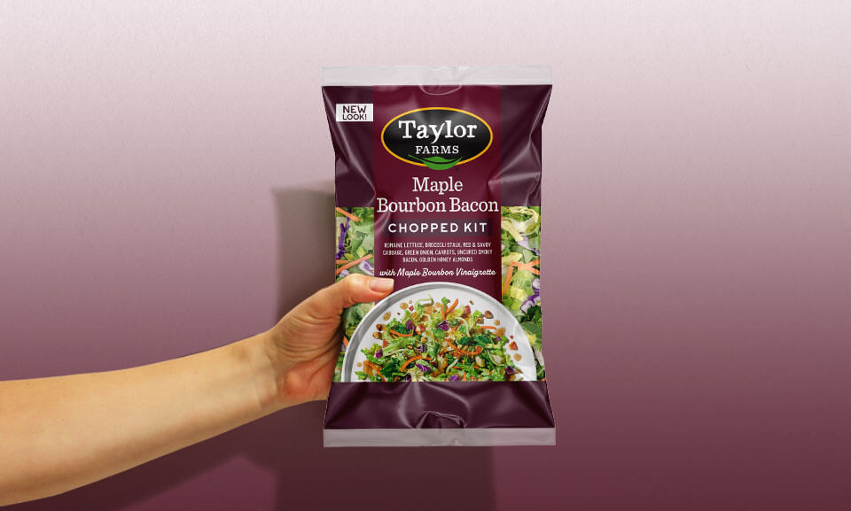 Hand holding a Maple Bourbon Bacon Chopped Salad Kit bag from Taylor Farms, showcasing fresh ingredients and packaging.