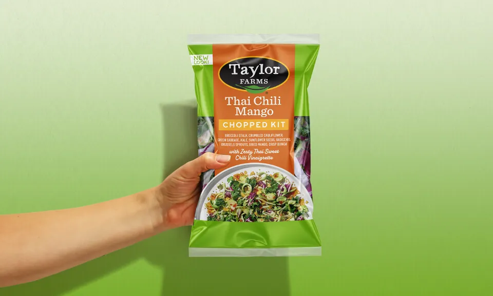 Hand holding a Thai Chili Mango Chopped Salad Kit bag from Taylor Farms, showcasing fresh ingredients and vibrant packaging.