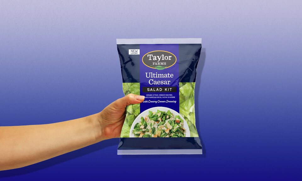 Hand holding a bag of Taylor Farms Ultimate Caesar Salad Kit, showcasing fresh ingredients and convenient packaging.