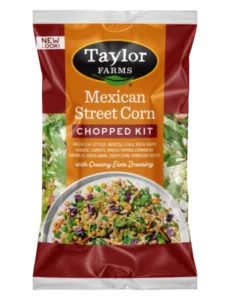 Taylor Farms Mexican Style Street Corn Chopped Salad Kit in a golden-colored pre-mixed package, showing the prepared salad with rows of corn, parmesan cheese, cornbread crumbles on a bed of green leaf lettuce with red cabbage and shredded carrots.