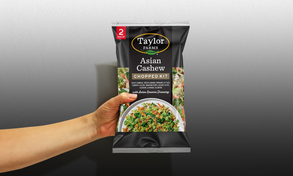 Hand holding a bag of Asian Cashew salad mix from Taylor Farms, showcasing fresh ingredients and vibrant packaging.